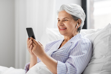 Poster - technology, old age and people concept - senior woman with smartphone and wireless earphones in bed at home bedroom