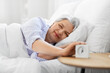 © Syda Productions - old age and people concept - happy smiling senior woman with alarm clock lying in bed at home bedroom