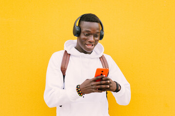 Wall Mural - Young african American black man with surprise face looking at smart phone while listening music with wireless headphones. Yellow wall wearing a white sweatshirt and a backpack