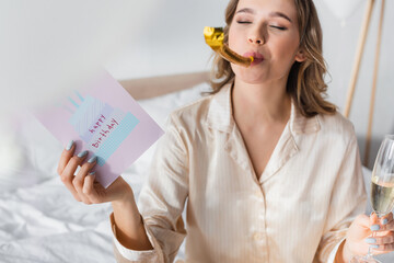 Birthday greeting card and champagne in hands of blurred woman with party horn in bedroom