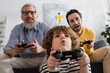© LIGHTFIELD STUDIOS - KYIV, UKRAINE - APRIL 12, 2021: Low angle view of excited child playing video game near father and granddad on blurred background