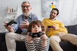 © LIGHTFIELD STUDIOS - KYIV, UKRAINE - APRIL 12, 2021: Astonished kid playing video game with blurred father and grandfather