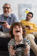 © LIGHTFIELD STUDIOS - KYIV, UKRAINE - APRIL 12, 2021: Excited kid playing video game near blurred parents