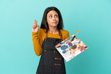 Young artist caucasian woman holding a palette isolated on blue background with fingers crossing and wishing the best