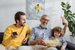 © LIGHTFIELD STUDIOS - Smiling man with popcorn waving hand near son with remote controller and grandson on couch