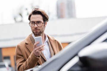 Wall Mural - confident man in glasses and beige coat holding mobile phone near blurred car