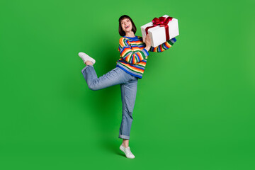 Poster - Full length photo of funny carefree young woman wear striped sweater smiling giving gift box isolated green color background