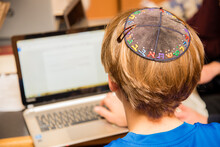 Young Jewish Boy Wearing Colorful Hebrew Yarmulke From The Back Typing On A Keyboard At School.