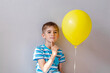 A pensive little boy in a blue striped T-shirt holds a yellow helium balloon on a gray neutral background. The child is thinking about something.