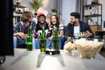 Wall Mural - Cheerful young people sitting on couch and using wireless microphone for singing in karaoke. Multiracial men and women spending weekends together with fun and pleasure.