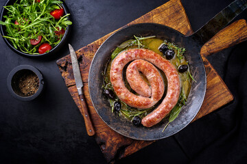 Wall Mural - Traditional fried Italian salsiccia fresco meat sausage served with rocket salad and olives as top view in a cast iron pan