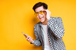 Cheerful asian guy in bright glasses holds a phone in his hand and laughs while standing on a yellow background