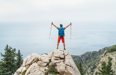 Wall Mural - young hiker backpacker man rose trekking poles up enjoying the mediterranean sea on clifftop during 