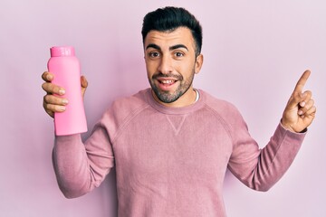 Wall Mural - Young hispanic man holding shampoo bottle smiling happy pointing with hand and finger to the side