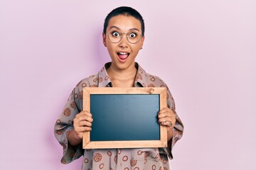 Wall Mural - Beautiful hispanic woman with short hair holding blackboard afraid and shocked with surprise and amazed expression, fear and excited face.