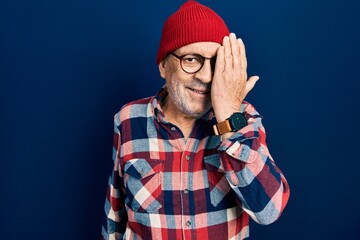 Wall Mural - Handsome mature man wearing hipster look with wool cap covering one eye with hand, confident smile on face and surprise emotion.