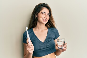 Poster - Young caucasian woman holding electric toothbrush and water glass winking looking at the camera with sexy expression, cheerful and happy face.