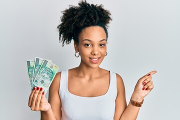 Poster - Young african american girl holding 100 polish zloty banknotes smiling happy pointing with hand and finger to the side