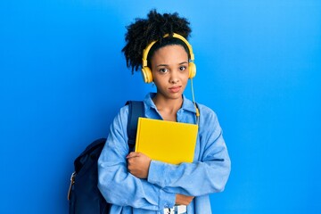 Wall Mural - Young african american girl wearing student backpack and headphones holding book clueless and confused expression. doubt concept.