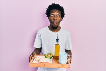 Poster - Young african american man holding tray with breakfast food making fish face with mouth and squinting eyes, crazy and comical.