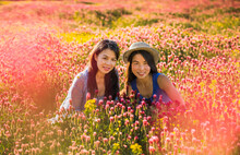 Two Middle-aged Chinese Women Friends Sitting In Meadow With Colorful Wildflowers And Smiling At Camera; Spring In Midwest