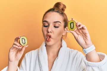 Poster - Young blonde woman wearing bathrobe holding fresh kiwi making fish face with mouth and squinting eyes, crazy and comical.