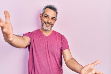 Poster - Handsome middle age man with grey hair wearing casual pink t shirt looking at the camera smiling with open arms for hug. cheerful expression embracing happiness.