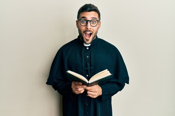 Young hispanic priest man holding bible celebrating crazy and amazed for success with open eyes screaming excited.