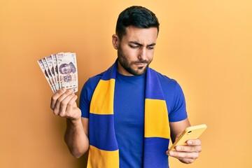 Poster - Young hispanic man football supporter using smartphone holding mexican pesos banknotes skeptic and nervous, frowning upset because of problem. negative person.