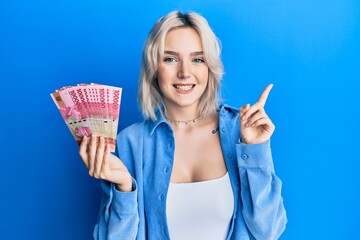 Sticker - Young blonde girl holding indonesian rupiah banknotes smiling happy pointing with hand and finger to the side