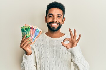 Wall Mural - Handsome hispanic man with beard holding south african rand banknotes doing ok sign with fingers, smiling friendly gesturing excellent symbol