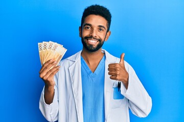 Canvas Print - Handsome hispanic man with beard wearing medical uniform holding 500 norwegian krone smiling happy and positive, thumb up doing excellent and approval sign