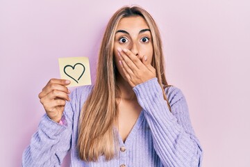 Canvas Print - Beautiful hispanic woman holding heart reminder covering mouth with hand, shocked and afraid for mistake. surprised expression