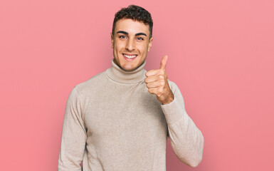 Hispanic young man wearing casual turtleneck sweater smiling happy and positive, thumb up doing excellent and approval sign