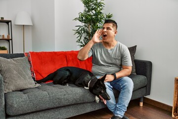 Sticker - Young latin man and dog sitting on the sofa at home shouting and screaming loud to side with hand on mouth. communication concept.