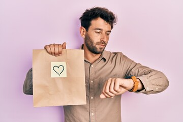 Poster - Handsome man with beard holding delivery paper bag with heart reminder checking the time on wrist watch, relaxed and confident