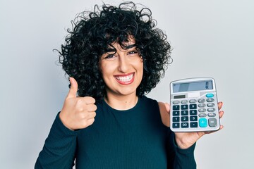 Sticker - Young middle east woman showing calculator device smiling happy and positive, thumb up doing excellent and approval sign