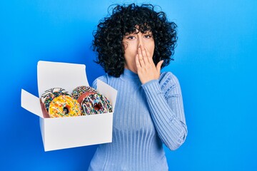 Poster - Young middle east woman holding tasty colorful doughnuts box covering mouth with hand, shocked and afraid for mistake. surprised expression