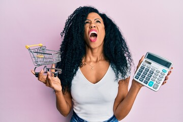 Middle age african american woman holding small supermarket shopping cart and calculator angry and mad screaming frustrated and furious, shouting with anger looking up.