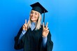 © Krakenimages.com - Beautiful blonde woman wearing graduation cap and ceremony robe smiling looking to the camera showing fingers doing victory sign. number two.