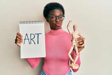 Canvas Print - Young african american girl holding wooden manikin and art word on notebook looking at the camera blowing a kiss being lovely and sexy. love expression.