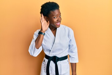 Canvas Print - Young african american girl wearing karate kimono and black belt smiling with hand over ear listening an hearing to rumor or gossip. deafness concept.