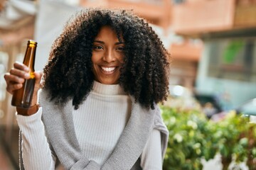 Poster - Young african american woman smiling happy holding bottle of beer at the city.