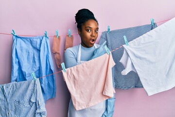 Poster - African american woman with braided hair washing clothes at clothesline surprised pointing with finger to the side, open mouth amazed expression.