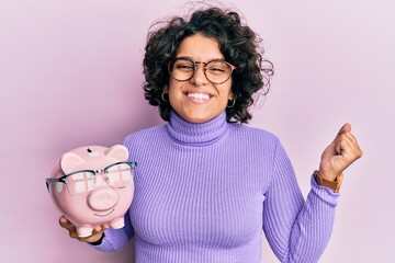 Young hispanic woman with curly hair holding piggy bank with glasses screaming proud, celebrating victory and success very excited with raised arm