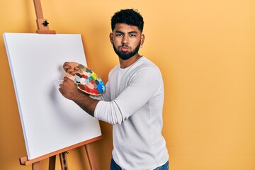 Sticker - Arab man with beard standing drawing with palette by painter easel stand puffing cheeks with funny face. mouth inflated with air, catching air.