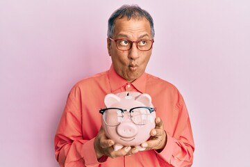 Sticker - Middle age indian man holding piggy bank with glasses making fish face with mouth and squinting eyes, crazy and comical.