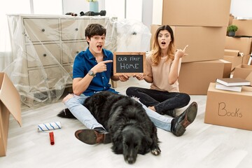 Poster - Young caucasian couple with dog holding our first home blackboard at new house surprised pointing with finger to the side, open mouth amazed expression.