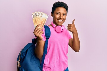 Wall Mural - Young african american girl wearing student backpack holding norwegian krone banknotes smiling happy and positive, thumb up doing excellent and approval sign