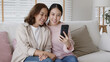 © ChayTee - Retired healthy mom and girl two people hold phone waving hand greeting selfie look at camera on webcam app at relax comfort sofa couch living room in health care quarantine social distance isolate.